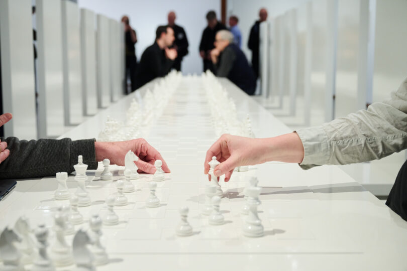 Foto von zwei Armen über einem unüblichen Schachbrett: In dem Kunstwerk „Play it by Trust“, 1966/2025, von Yoko Ono in der Neuen Nationalgalerie sind Besuchende aufgefordert, mit rein weißen Figuren Schach zu spielen.