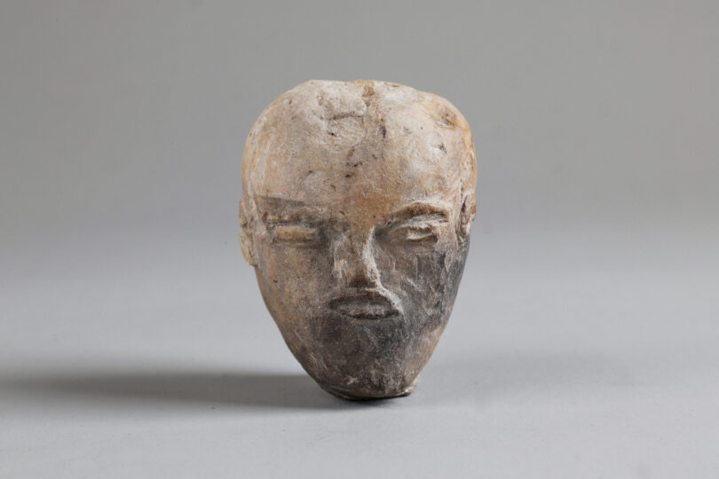 Das Bild zeigt einen aus Kalkstein gefertigten Menschenkopf. Die Gesichtszüge sind klar erkennbar; die Augen wirken zusammengekniffen. Der Kopf ist unbehaart.