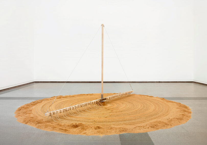 Das Bild zeigt einen Kreis aus Sand auf dem Boden. Darüber befindet sich ein Holzaufbau, der sich im Kreis dreht und den Sand mithilfe von Fäden glatt zieht.