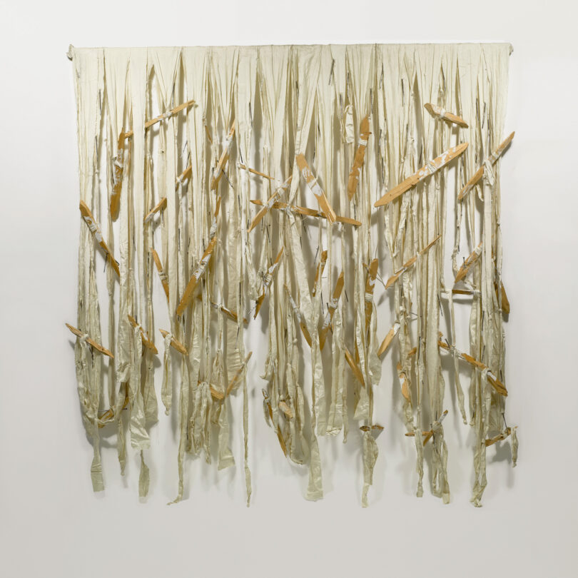 Auf dem Bild sind weiße Stofffäden zu sehen, die dicht nebeneinander hängen. An einigen Fäden sind Holzstäbchen angebunden.