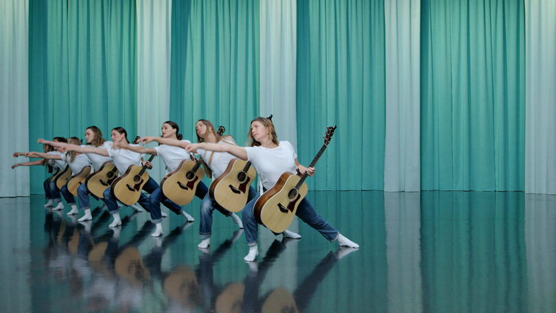 Auf dem Foto sieht man Tänzerinnen der Iceland Dance Company, die Gitarre spielen. Im Hintergrund hängen türkisfarbene Vorhänge