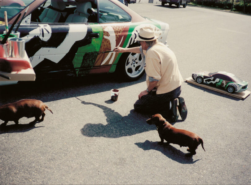David Hockney bemalt das 14. BMW Art Car. Neben ihm sind zwei Dackel zu sehen
