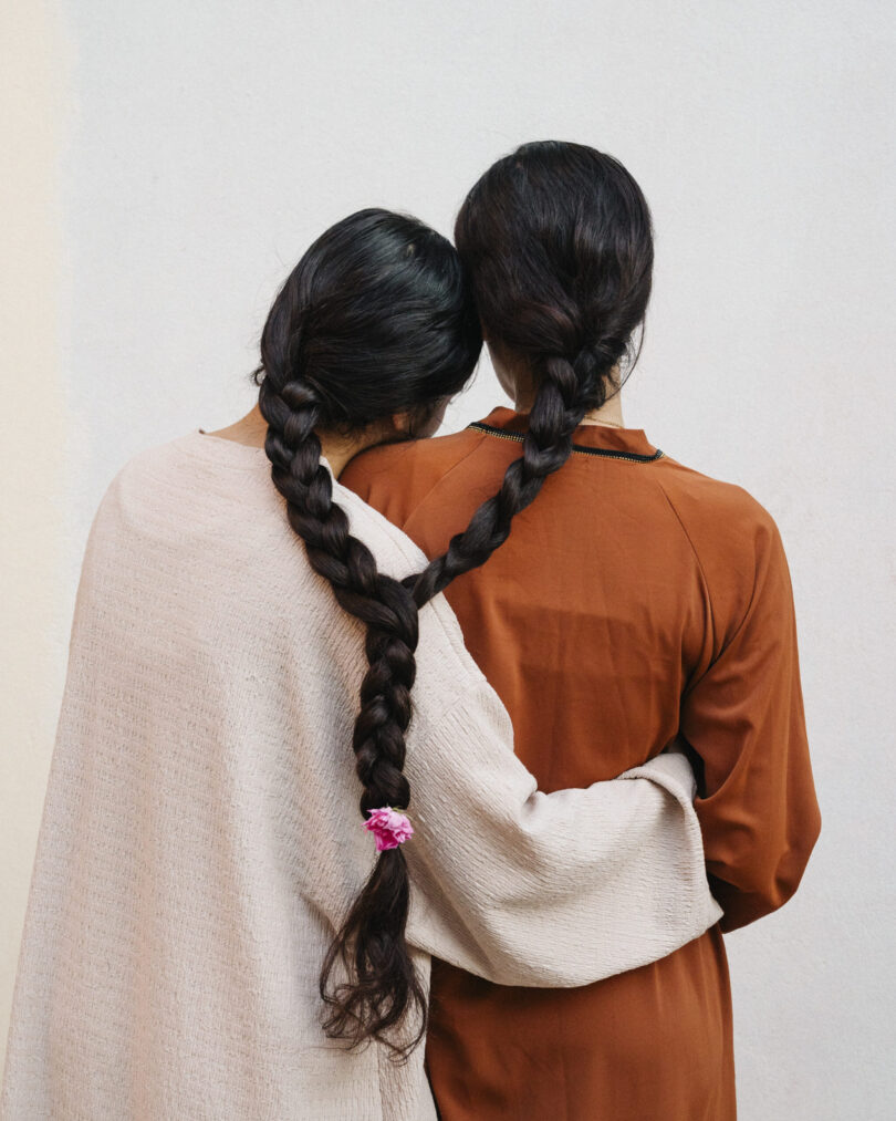Zwei Frauen stehen mit dem Rücken zum Betrachter, ihre langen schwarzen Haare sind zu einem gemeinsamen Zopf geflochten.