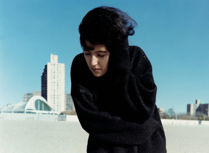 Fotografie einer in schwarz gekleideten Frau, die mit geschlossenen Augen und Wind in den Haaren im Freien vor strahlend blauem Himmel steht.